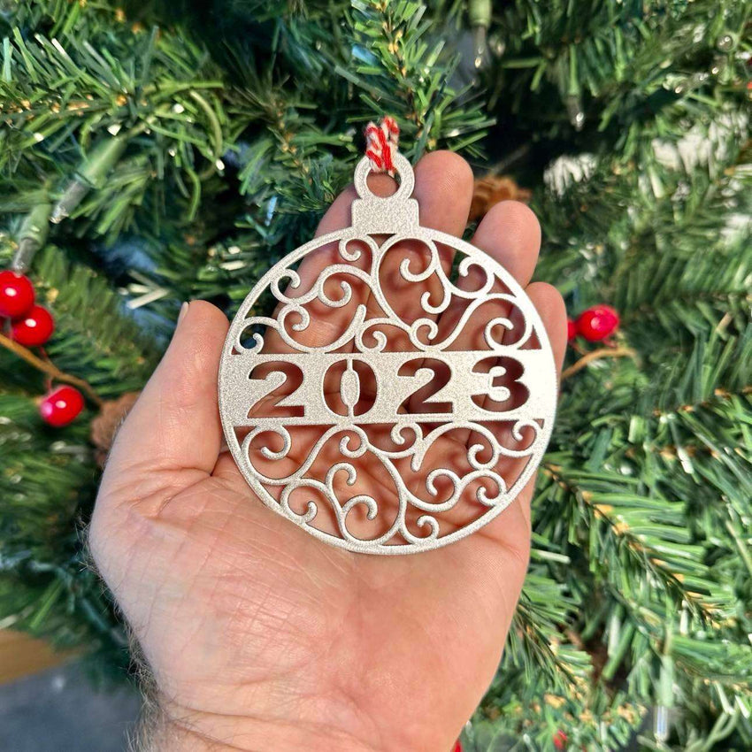 Decorative Christmas ornament displaying “2023” held in front of a Christmas tree, with soft green branches and festive lights in the background creating a warm holiday atmosphere.