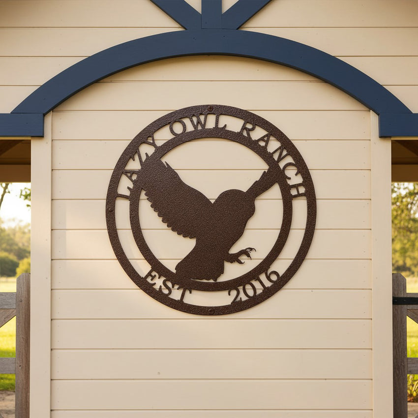 A round, laser-cut steel sign featuring a textured hammered bronze finish. The center design showcases the silhouette of an owl mid-flight with its wings spread, framed by a circular border containing the words "LAZY OWL RANCH" on top and "EST. 2016" along the bottom. The sign is mounted on a light cream horizontal lap siding accent wall under a decorative dark blue wooden archway.