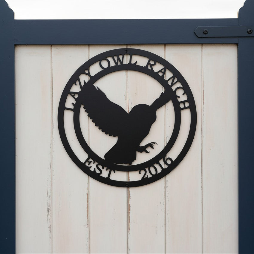A circular matte or satin black metal property sign depicting a flying owl silhouette, bordered by the custom inscriptions "LAZY OWL RANCH" and "EST. 2016". The sign is mounted on the white vertical wood paneling of a garden gate framed by deep navy blue posts.