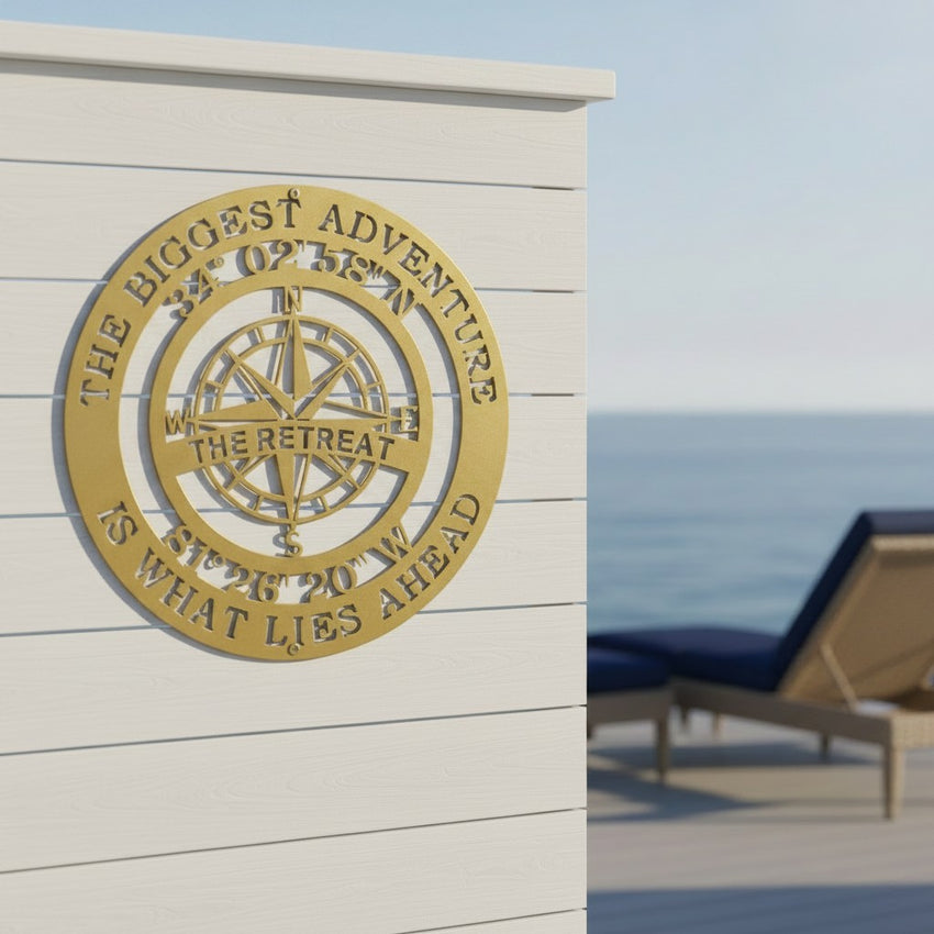 An elegant gold circular metal cut-out sign depicting an intricate compass rose is mounted on a clean white horizontal shiplap wall. The sign features custom text reading "THE RETREAT" and global coordinates. An outdoor seaside terrace with a blue deck chair is visible to the right.