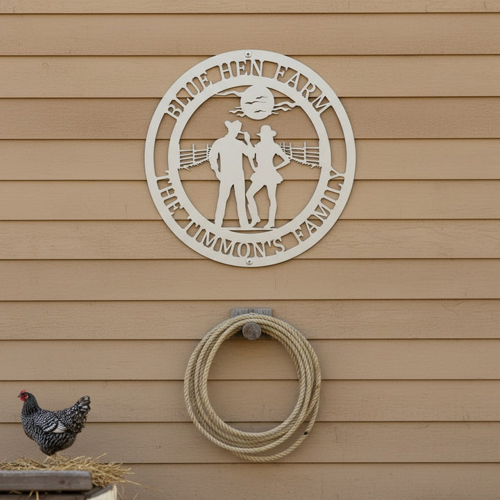 White personalized metal farm sign showing a cowboy and cowgirl silhouette, mounted on tan horizontal siding above a coiled rope with a speckled chicken nearby.
