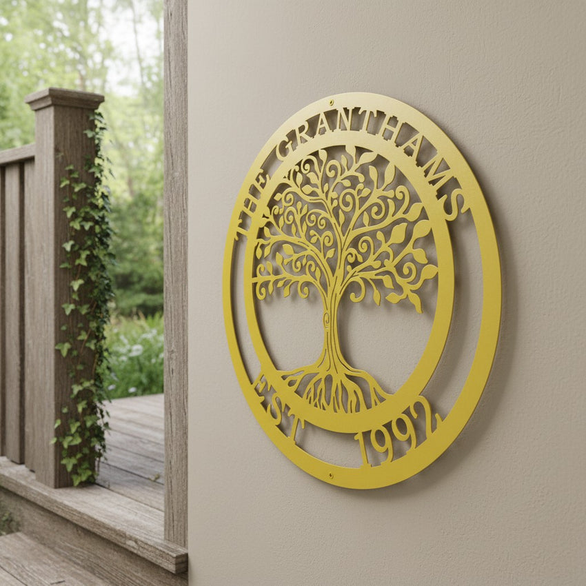 A bright mustard yellow circular metal cutout sign customized with the text "THE GRANTHAMS EST. 1992" around a central tree motif. The piece is hanging on a beige exterior wall column beside a wooden porch railing wrapped in climbing green ivy.