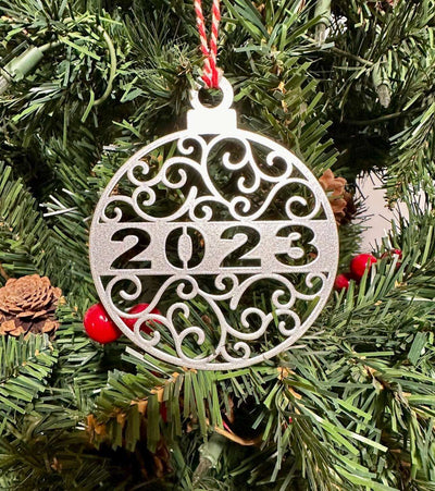 Decorative Christmas ornament displaying “2023” hanging on a tree, surrounded by green branches and festive holiday decorations, creating a cheerful and seasonal atmosphere.