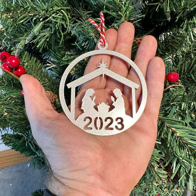 Decorative Christmas ornament displaying “2023” held in a hand in front of a Christmas tree, with green branches and festive lights in the background creating a warm holiday scene.