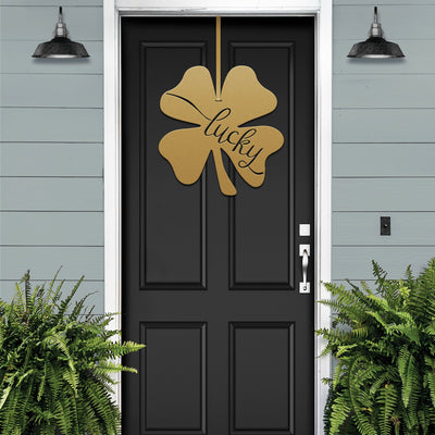 Lucky Shamrocks metal door wreath with gold four-leaf clover design and “Lucky” text on black door, festive Irish décor with plants on light blue wall.