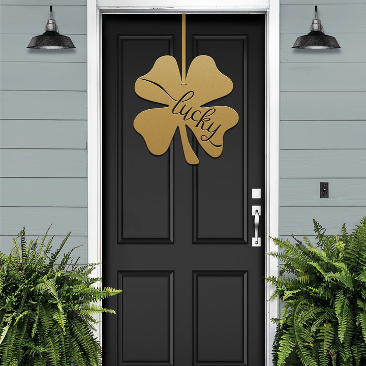 Lucky Shamrocks metal door wreath with gold four-leaf clover design and “Lucky” text on black door, festive Irish décor with plants on light blue wall.