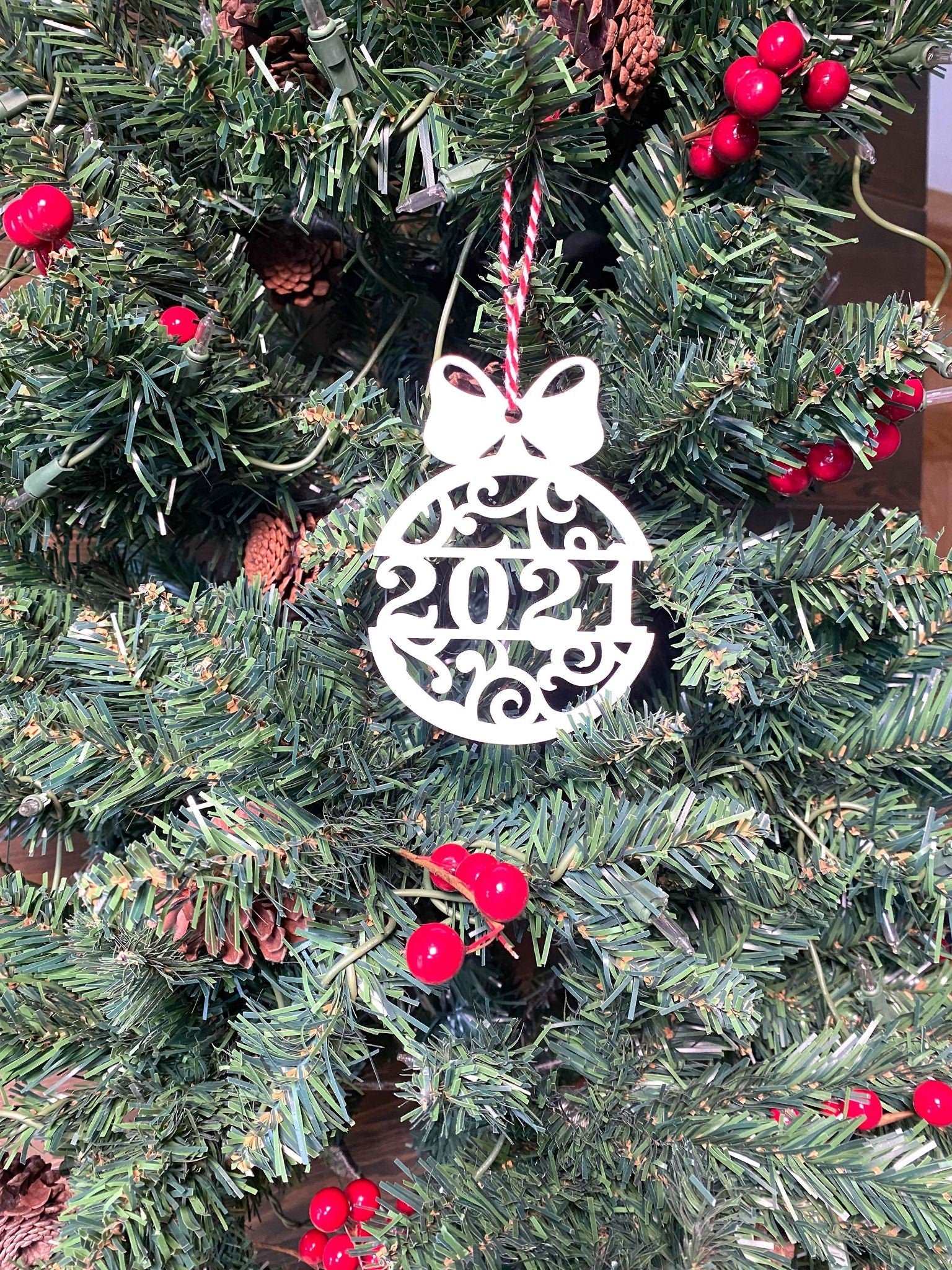 White 2021 scroll Christmas ball ornament hanging on green holiday tree with red berries and pinecones