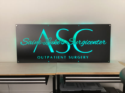 Custom matte black LED backlit metal sign for Saint Luke’s Surgicenter ASC Outpatient Surgery, blue illuminated text on powder coated steel wall signage, modern medical office branding sign for indoor lobby or front door outdoor entry display