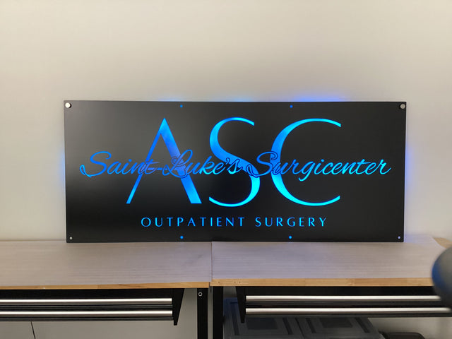 Custom matte black LED backlit metal sign for Saint Luke’s Surgicenter Outpatient Surgery, blue illuminated text on powder coated steel wall sign, modern medical office branding signage for indoor lobby or outdoor entry display