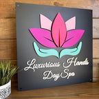 Custom 3D multi-material steel metal sign with pink and green lotus flower design and "Luxurious Hands Day Spa" text mounted on a wooden background – House Sensations Art.