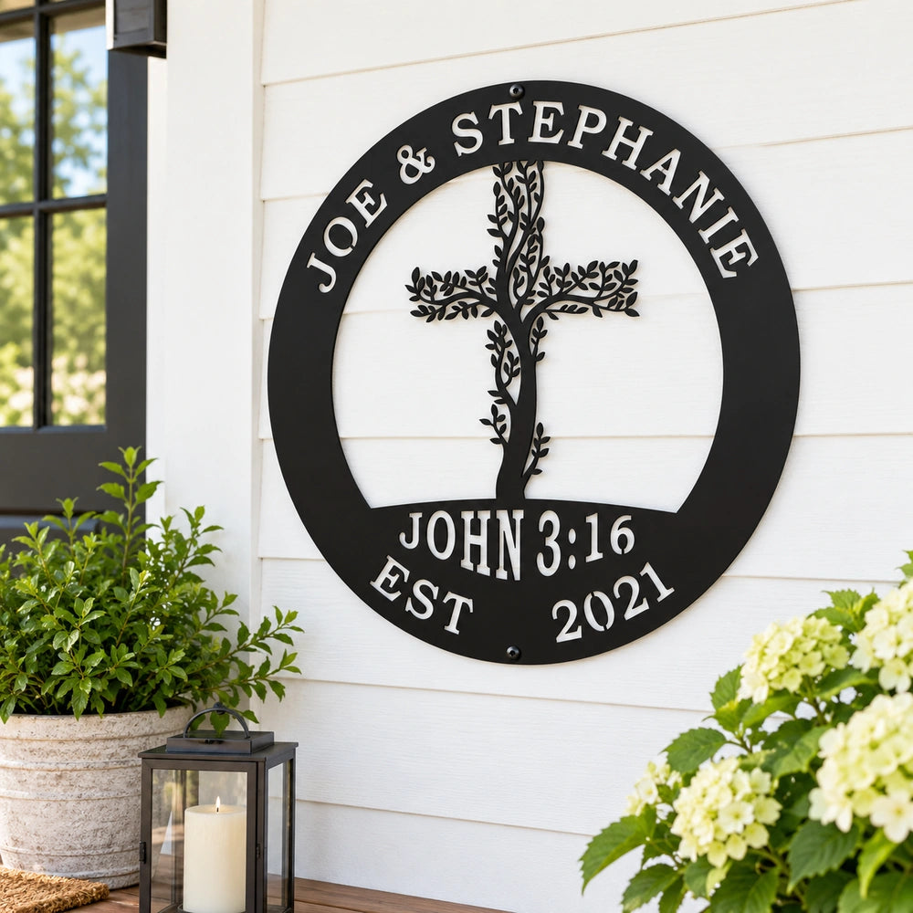 A black metal sign with a tree-like design and the names "Joe & Stephanie" is mounted on a white wall, with a potted plant and a lantern to the left and a plant with white flowers to the right.