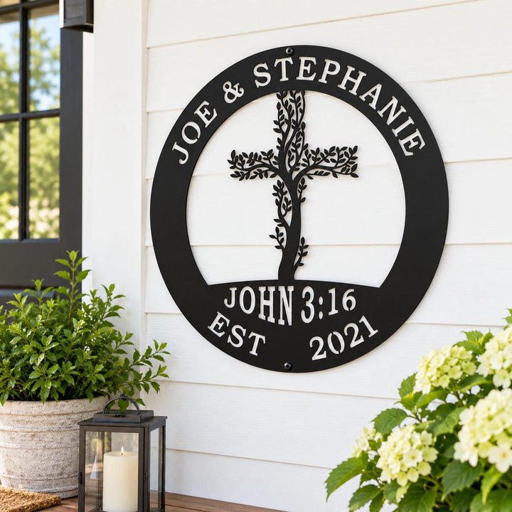 A black metal sign with a tree-like design and the names "Joe & Stephanie" is mounted on a white wall, with a potted plant and a lantern to the left and a plant with white flowers to the right.
