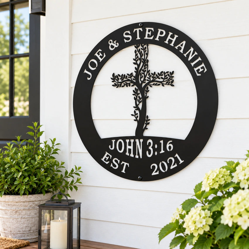 A black metal sign with a tree-like design and the names "Joe & Stephanie" is mounted on a white wall, with a potted plant and a lantern to the left and a plant with white flowers to the right.
