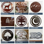 9-panel finish and color chart for custom metal signs featuring earthy and metallic tones. Top row includes Tuscan Copper dog-themed sign, Candy Copper personalized windmill sign, and Textured Copper mountain elevation sign. Middle row shows Espresso logo plaque, Hammered Bronze tree farm sign, and Oil Rubbed Bronze moon-and-stars camping sign. Bottom row features Black Bronze oak tree sign, Satin Titanium cabin and bear welcome sign, and Satin Silver monogram plaque.