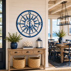 A round custom metal wall plaque finished in a deep coastal blue, highlighting an anchor integrated into a large compass rose. The sign is centered on a light-colored horizontal plank interior wall directly above a rustic wooden console table in a bright beach-themed dining room.