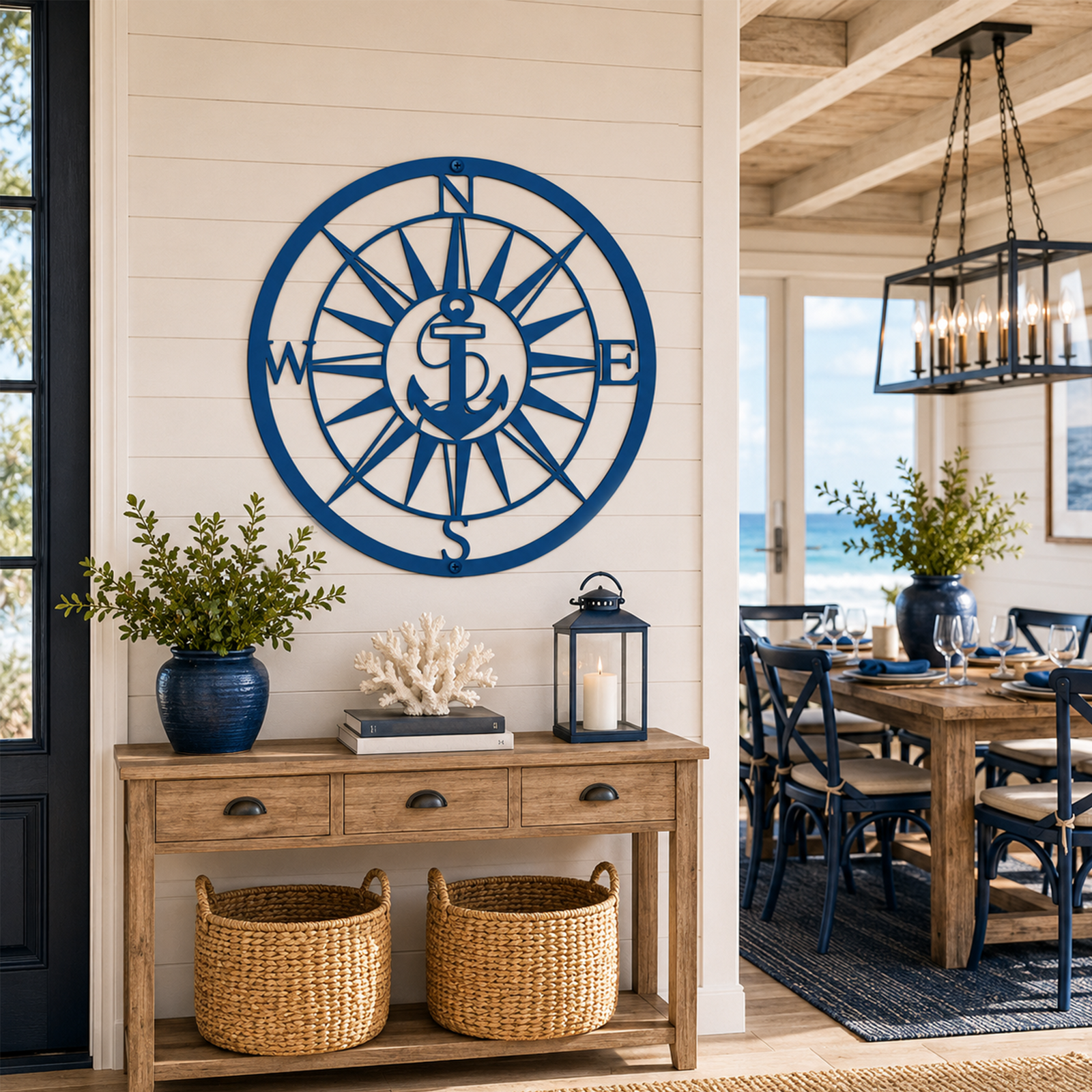 A round custom metal wall plaque finished in a deep coastal blue, highlighting an anchor integrated into a large compass rose. The sign is centered on a light-colored horizontal plank interior wall directly above a rustic wooden console table in a bright beach-themed dining room.