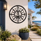 A round, laser-cut matte black metal wall sign featuring a classic anchor nested in the center of an intricate compass rose with N, S, E, and W markers. The sign is mounted on a smooth cream stucco pillar next to a dark blue front door and a stone-lined walkway leading toward the ocean.