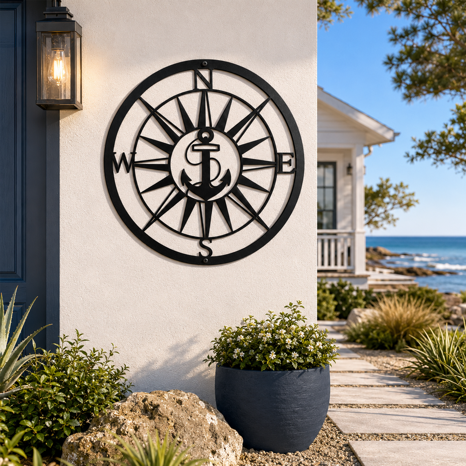 A round, laser-cut matte black metal wall sign featuring a classic anchor nested in the center of an intricate compass rose with N, S, E, and W markers. The sign is mounted on a smooth cream stucco pillar next to a dark blue front door and a stone-lined walkway leading toward the ocean.