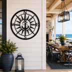 A round black metal anchor compass plaque mounted on the white vertical siding of an upscale outdoor veranda. The background features a long wooden family dining table set for guests, looking directly out onto blue ocean waves.