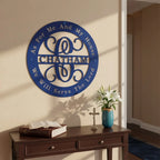 A circular royal blue metal monogram sign featuring the family name "CHATHAM" inside a decorative flourish centerpiece. The border is inscribed with the Bible verse, "As For Me And My House We Will Serve The Lord." The sign is mounted on a beige hallway wall above a dark wood console table decorated with an open Bible, a crucifix, and a blue vase with white lilies.