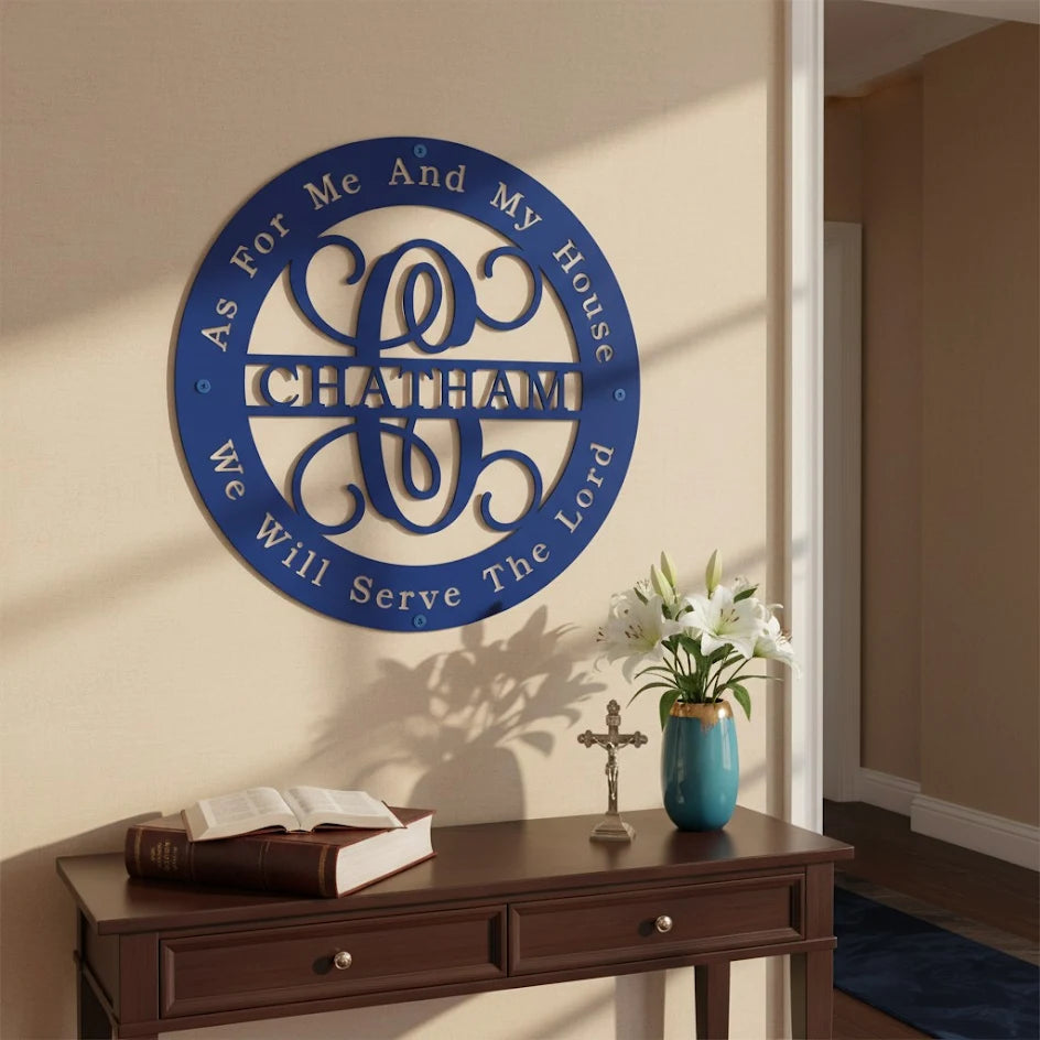 A circular royal blue metal monogram sign featuring the family name "CHATHAM" inside a decorative flourish centerpiece. The border is inscribed with the Bible verse, "As For Me And My House We Will Serve The Lord." The sign is mounted on a beige hallway wall above a dark wood console table decorated with an open Bible, a crucifix, and a blue vase with white lilies.
