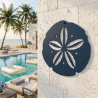 A dark navy blue metal sand dollar cutout decoration hanging on a white textured stone pillar, overlooking a luxury resort pool area with palm trees and a sandy beach.