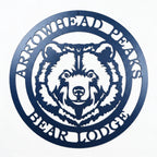 Custom Arrowhead Peaks Bear Lodge metal sign mounted above wooden shelf with books and decorative plants on white shiplap wall