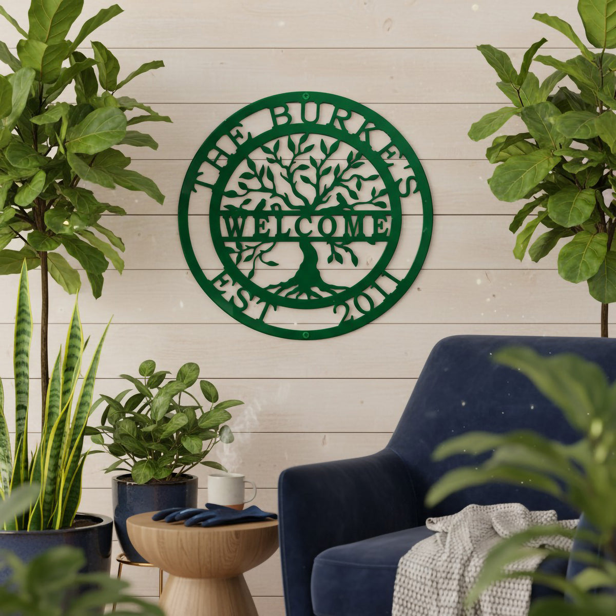 A forest green round metal custom sign with a family name and tree illustration, mounted on a white wood wall behind a blue armchair and flanked by large fiddle leaf fig plants.