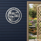  A silver metallic round custom sign reading "Jane's Pond Est. 2022" with a carved koi fish illustration, fixed to a dark blue horizontal siding wall next to an open doorway.
