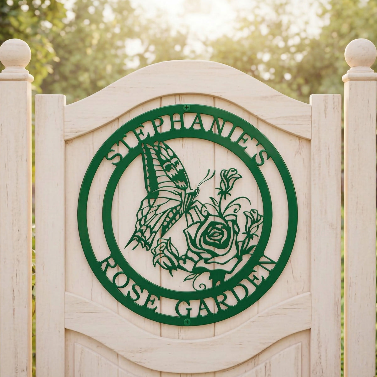A forest green circular metal sign reading "Stephanie's Rose Garden" with a carved butterfly motif, mounted centered on an arched white wooden garden fence.