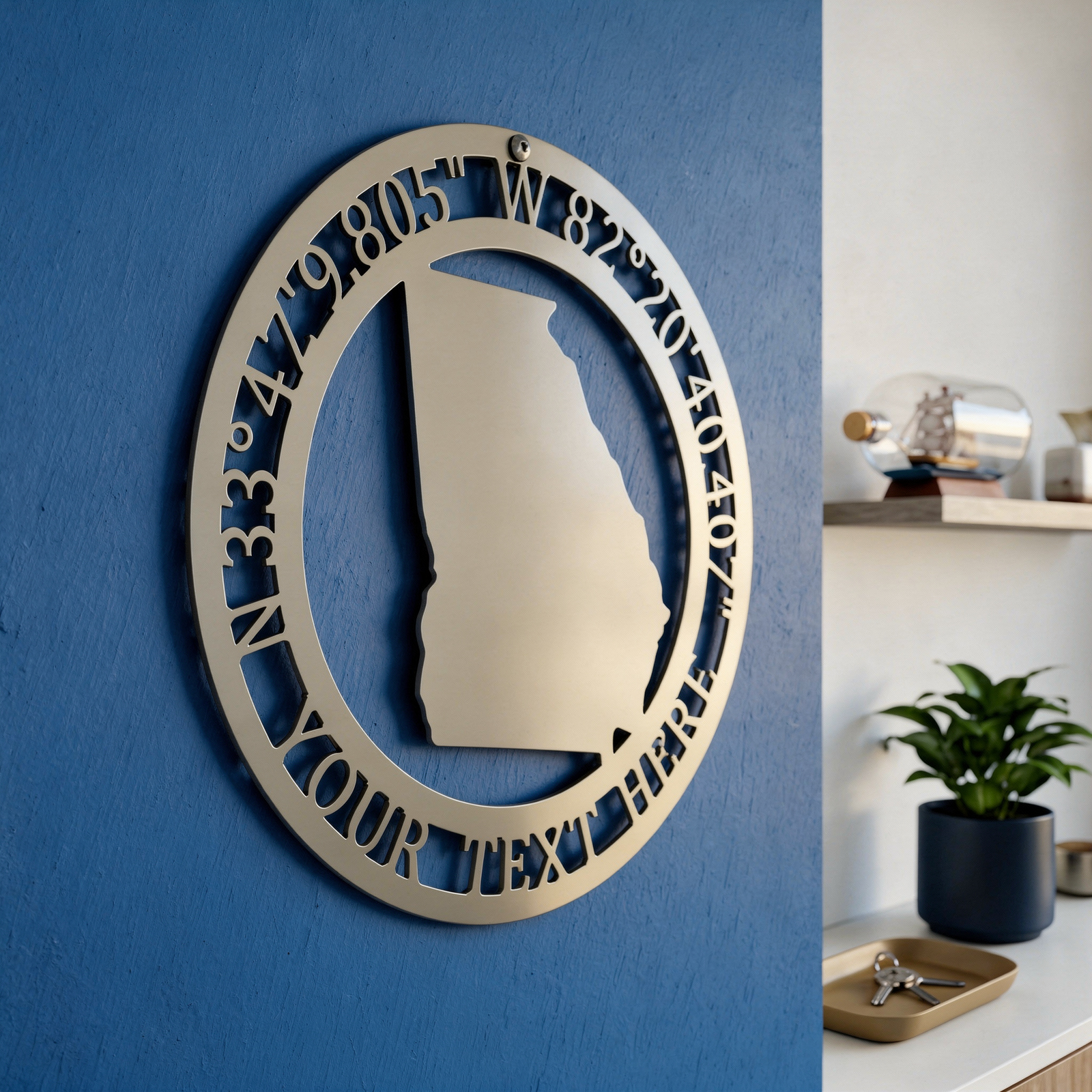A silver metallic circular sign featuring the outline of Georgia and latitude/longitude details, mounted at an angle on a deep blue textured wall next to an entryway shelf.
