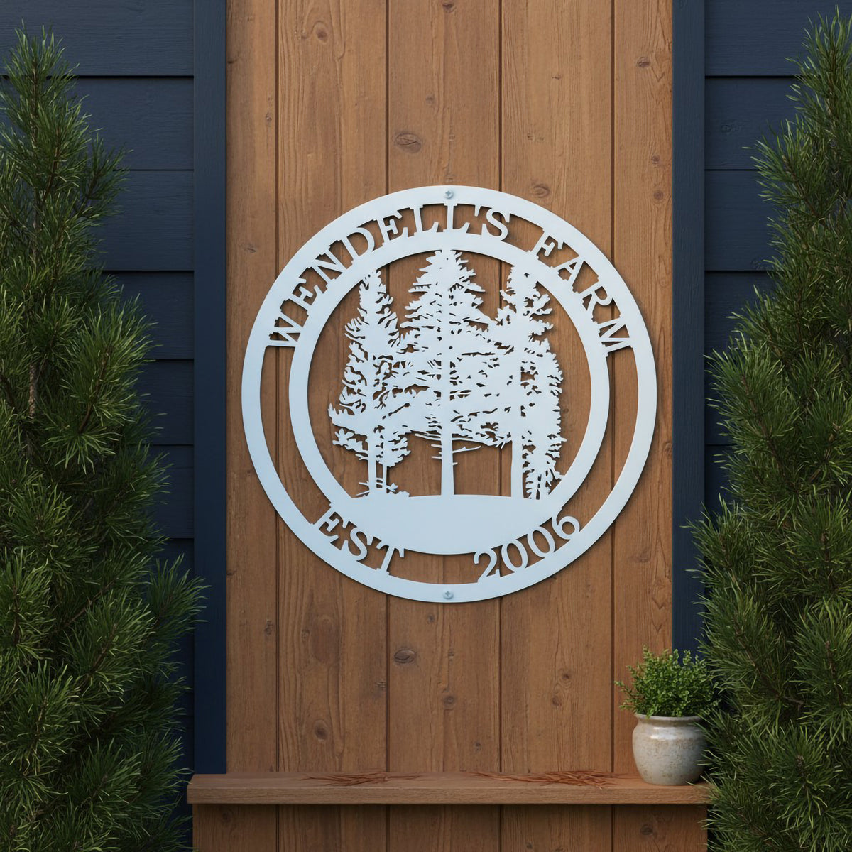  A white circular metal tree sign reading "Wendell's Farm Est. 2006," mounted on a vertical natural wood accent panel between two potted evergreen trees.