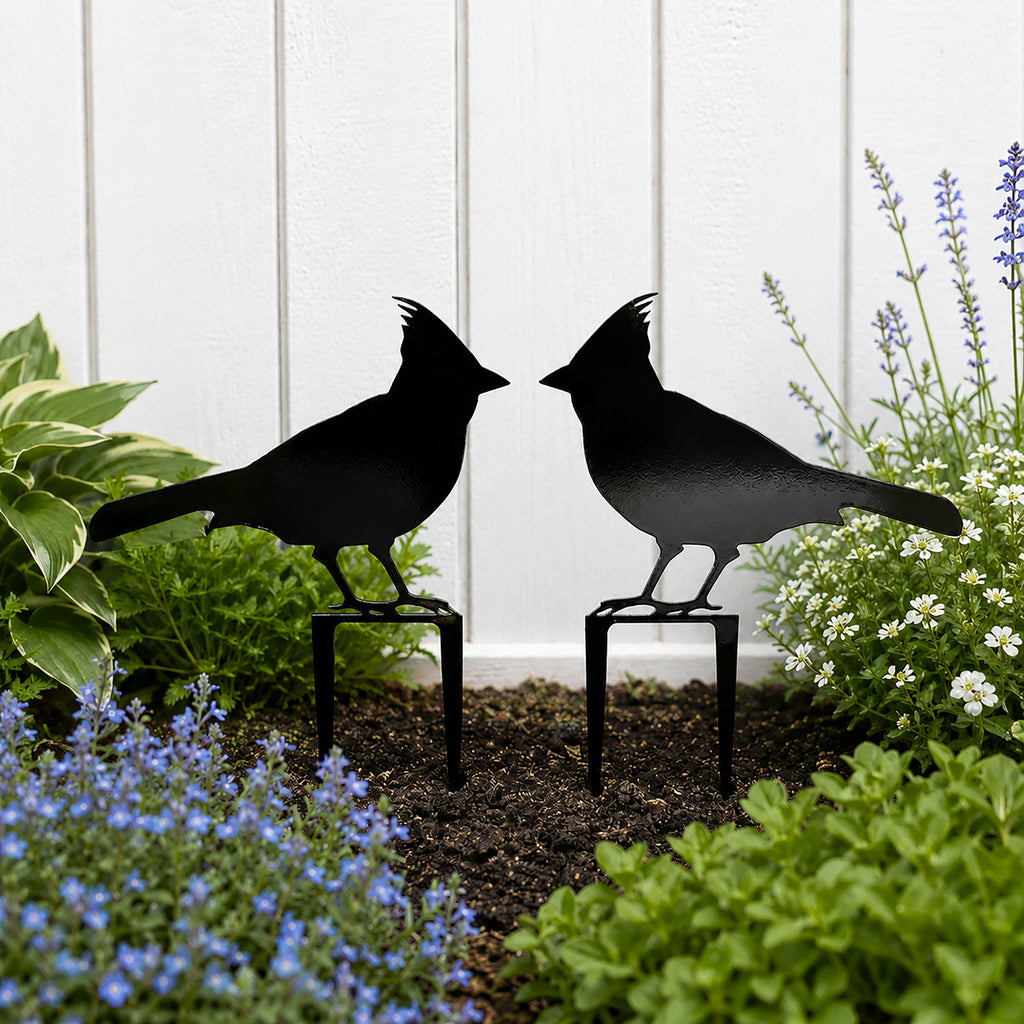 Black cardinal metal garden stakes displayed in a flower bed against a white fence with lush garden plants