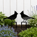 Black cardinal metal garden stakes displayed in a flower bed against a white fence with lush garden plants