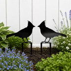 Black cardinal metal garden stakes displayed in a flower bed against a white fence with lush garden plants