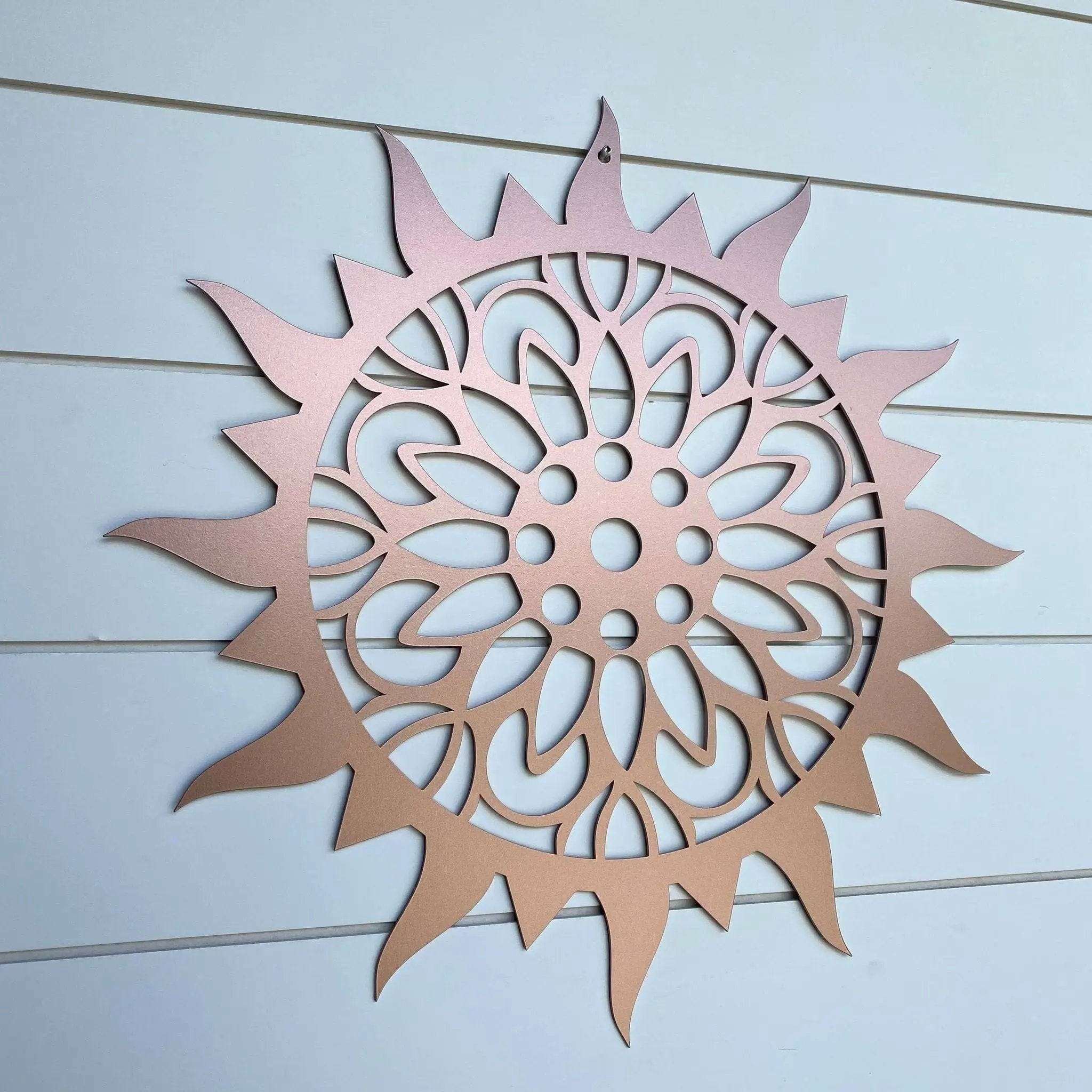 Blush pink sunburst mandala metal wall art featuring intricate floral cutout pattern mounted on white exterior siding for modern boho outdoor or indoor decor