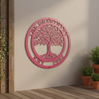 A vibrant bubblegum pink circular metal sign featuring a detailed Tree of Life cutout and the custom name "THE GRANTHAMS EST. 1992." The colorful monogram piece is mounted on an off-white wall above a wooden patio deck next to three stacked terracotta potted plants.