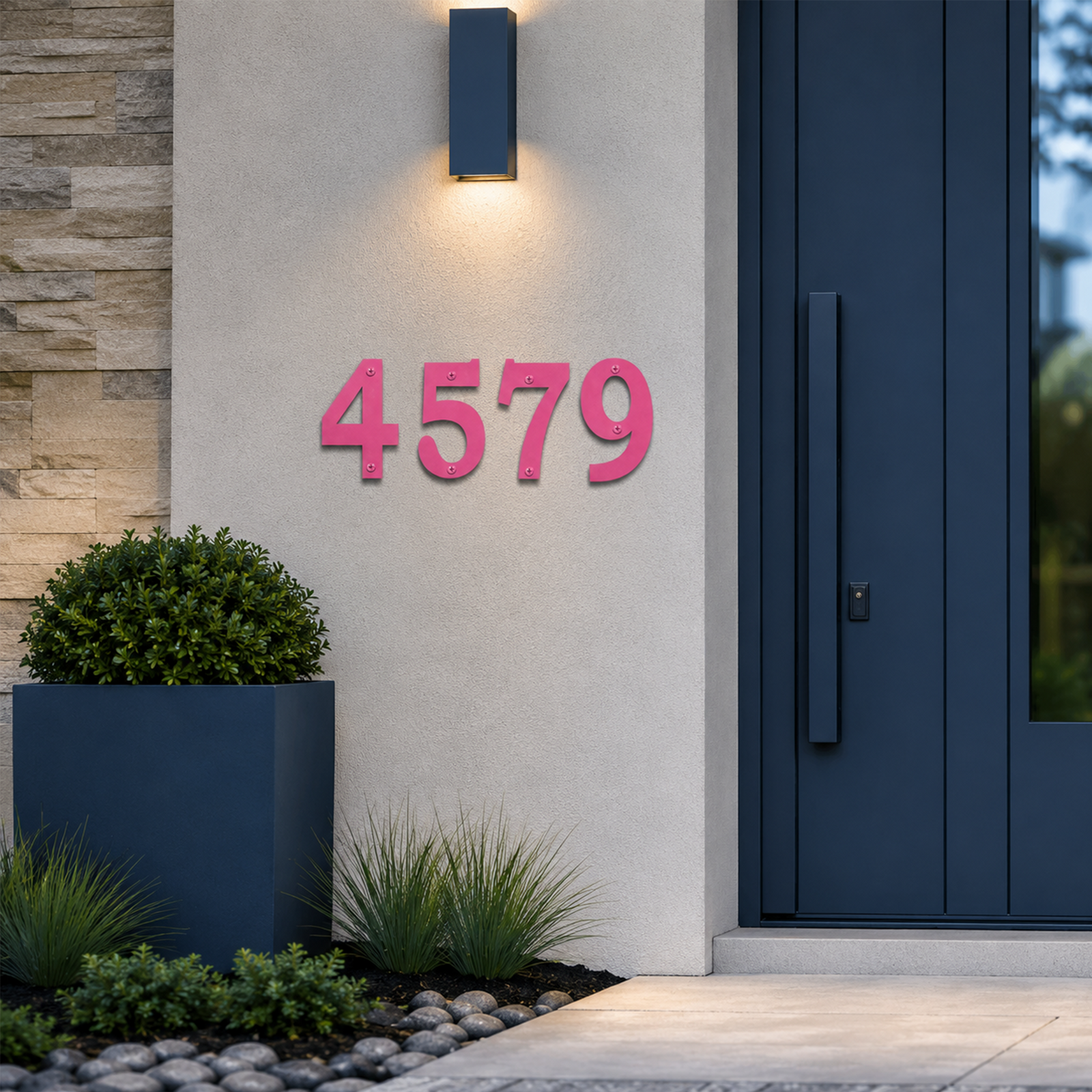 Eye-catching powder-coated hot pink metal house numbers displaying "4579" are mounted on a light concrete wall. The vibrant digits contrast sharply against a dark navy blue modern door frame and a stone accent wall panel, surrounded by minimalist landscaping with river stones and green grass.