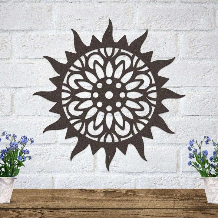 Intricate bronze sunburst metal wall art on white brick above a wooden shelf with small blue potted flowers.