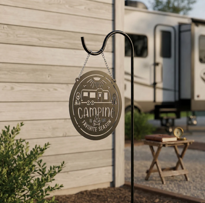 Brushed metal camping sign hanging from a black shepherd hook near an RV campsite, featuring camper trailer artwork, campfire details, and Camping Is Our Favorite Season text.