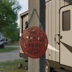 Deep red camping metal sign hanging from a wooden campsite post beside an RV trailer, featuring a camper trailer illustration and Camping Is Our Favorite Season wording.