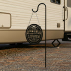 Gloss black camping metal sign suspended from a shepherd hook near an RV trailer featuring a camper, campfire, and Camping Is Our Favorite Season cutout design.