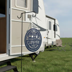 Navy blue camping metal sign hanging from a shepherd hook beside an RV campsite featuring a camper trailer and Camping Is Our Favorite Season design.