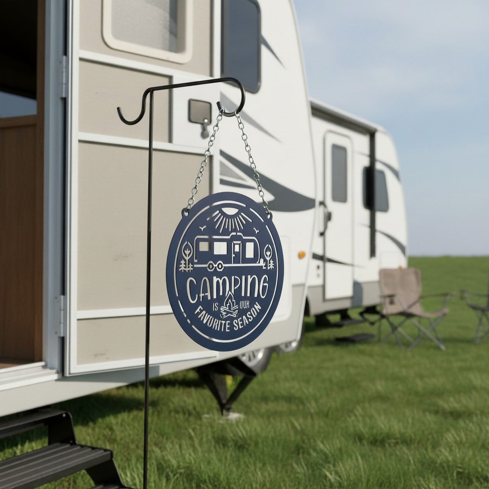 Navy blue camping metal sign hanging from a shepherd hook beside an RV campsite featuring a camper trailer and Camping Is Our Favorite Season design.