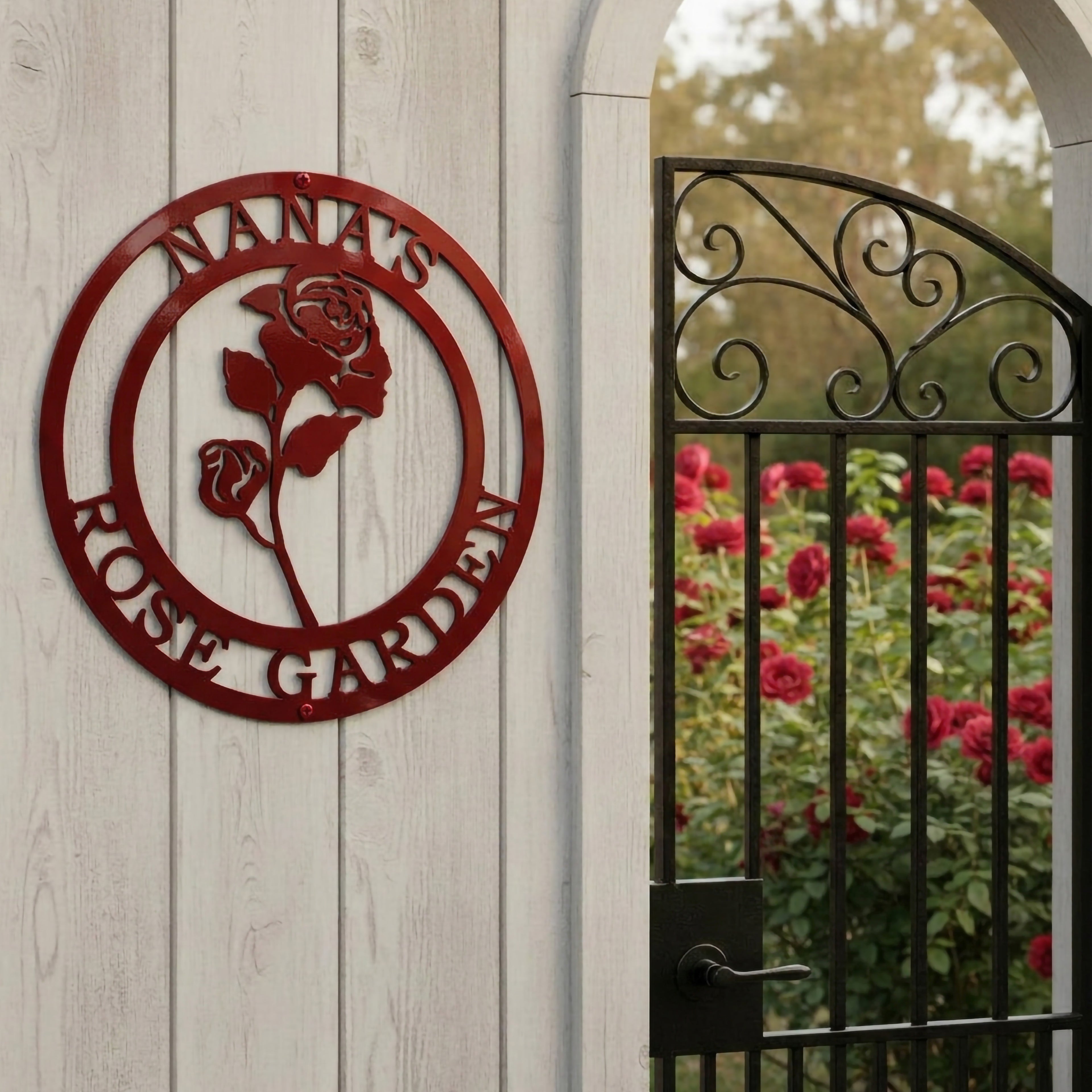 Glossy candy apple red finish of the custom circular rose metal art sign styled on a white vertical wood panel wall next to an open metal gate archway leading to a blooming rose field.
