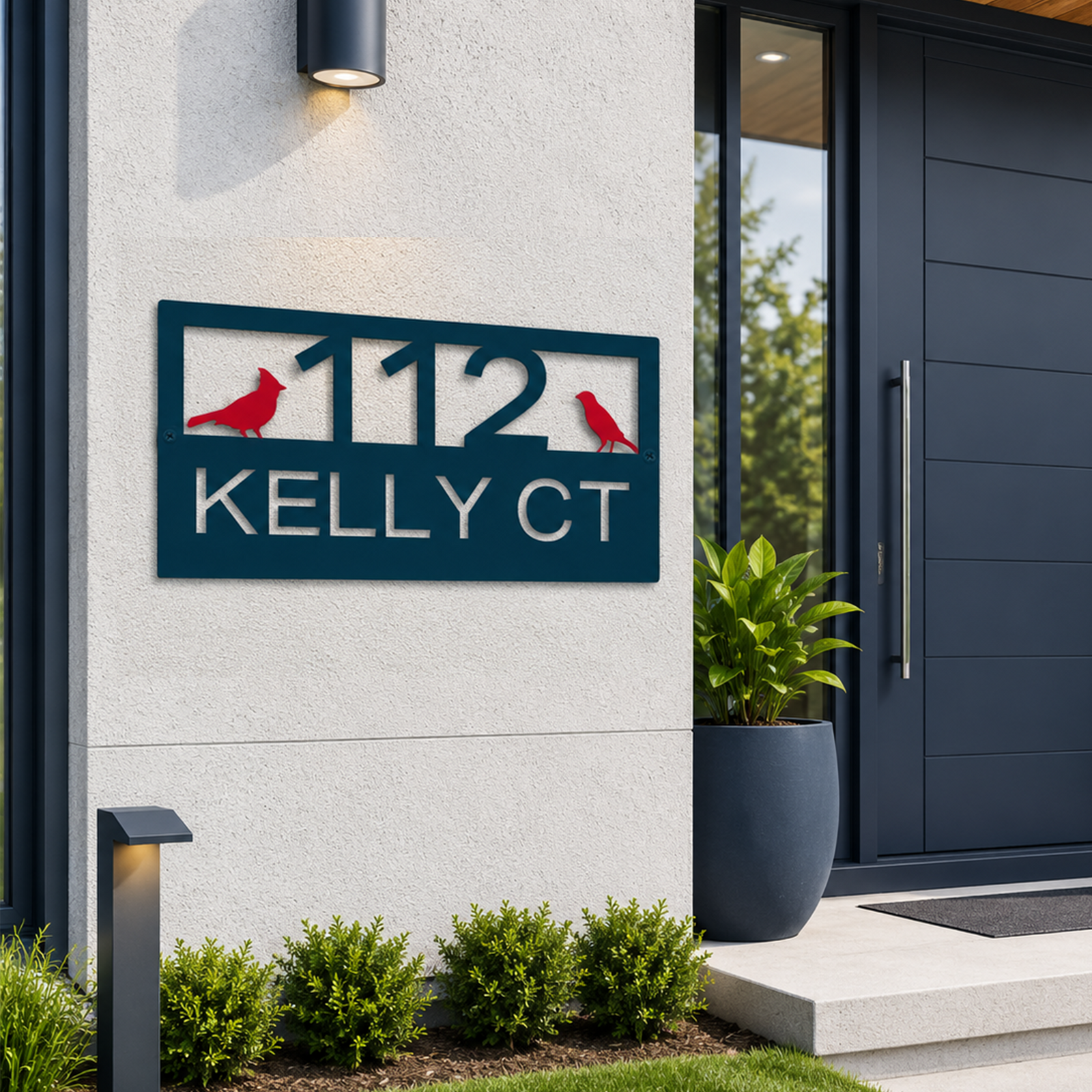 A rectangular metal address plaque in a deep ocean blue finish is mounted on a textured cream exterior wall. The sign displays the cutout text "112 KELLY CT" accented by two bright red cardinal bird silhouettes. A large navy planter sits by a matching modern front door in the background.