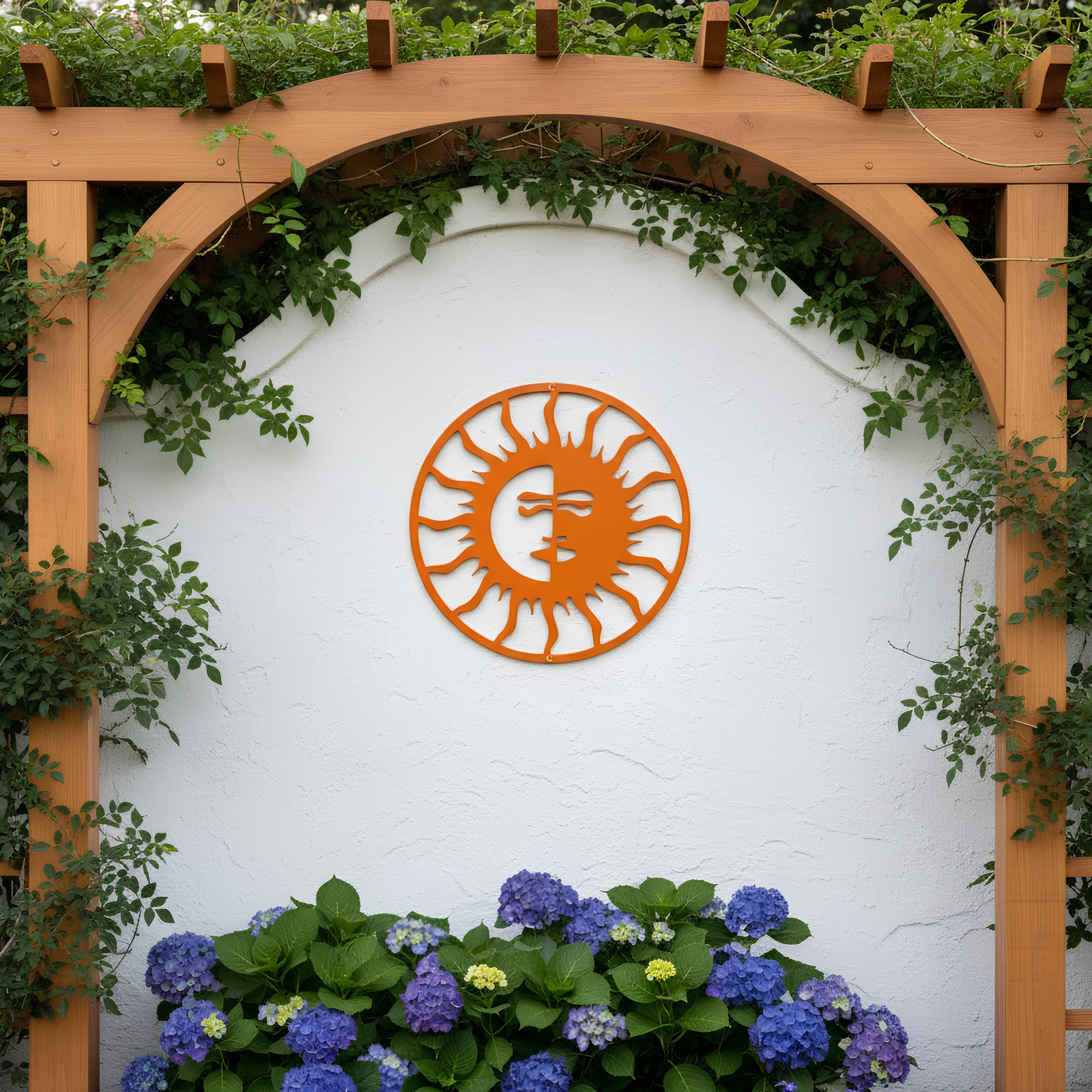 A vibrant orange circular metal sign featuring an intricate sun face design with wavy rays, mounted centrally on a white stucco wall underneath a wooden garden pergola flanked by blue hydrangeas.