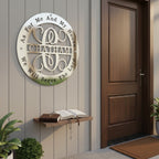 A satin silver circular metal plaque reading "CHATHAM" and engraved with the religious quote "As For Me And My House We Will Serve The Lord." The sign is installed on a light gray vertical siding porch wall next to a dark wood front door. An open Bible with a rosary rests on a small wooden shelf below it.