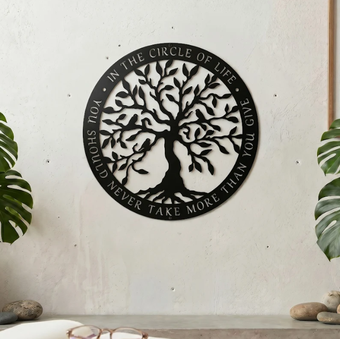 circular matte black metal sign featuring an intricate Tree of Life silhouette is mounted on a light beige exterior wall column. The outer border contains the cut-out quote, "IN THE CIRCLE OF LIFE, YOU SHOULD NEVER TAKE MORE THAN YOU GIVE." Lush green bamboo plants stand in the background on either side of a wooden door.