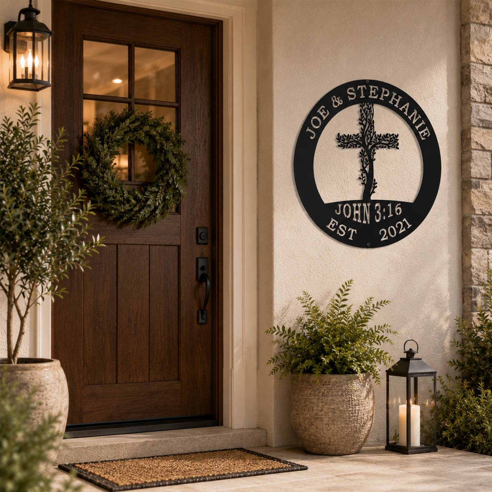 Black custom floral cross metal sign with the names Joe & Stephanie, hung outdoors on a white wall beside a dark wood front door with a green wreath.